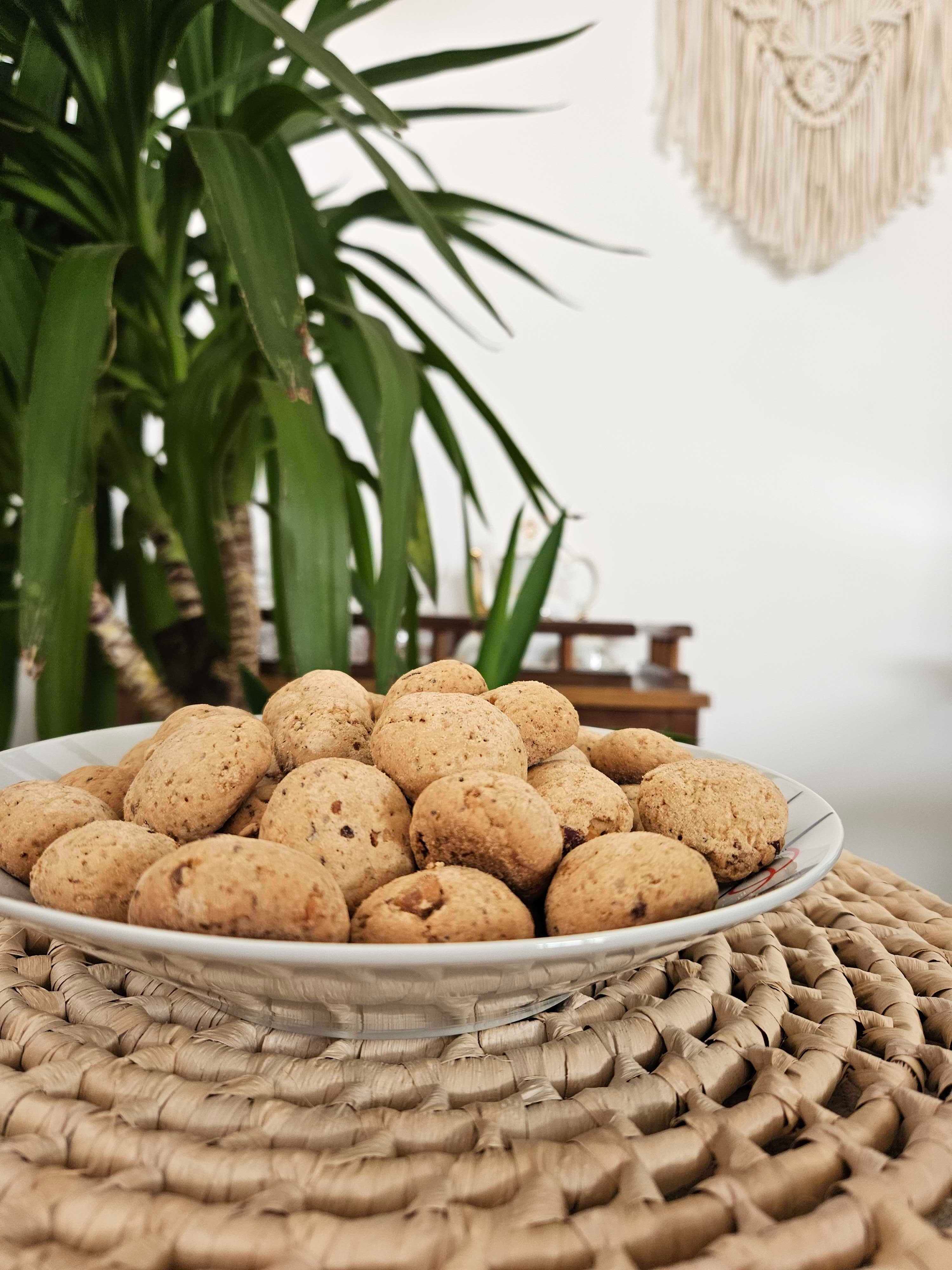 Prato de porcelana cheio de CadimBom, biscoito artesanal de amendoim sem glúten e sem lactose. Em mesa de vidro com plantas e uma xícara de café.